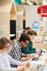 Group of concentrated multiethnic students sitting in line and doing hometask in library, focus on African guy typing on laptop