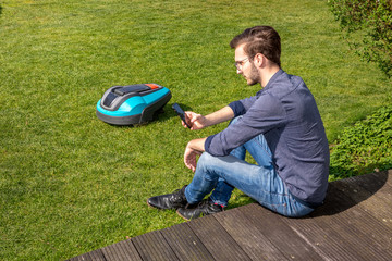 junger Mann sitzt mit Handy neben Roboterrasenmäher im Garten