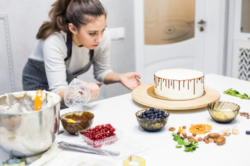 A confectioner squeezes liquid chocolate from a pastry bag onto a white cream biscuit cake on a wooden stand. The concept of homemade pastry, cooking cakes.