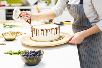 A confectioner squeezes liquid chocolate from a pastry bag onto a white cream biscuit cake on a wooden stand. The concept of homemade pastry, cooking cakes.