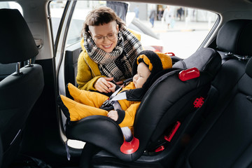A pretty brunette in a yellow jacket fastens the child car seat belt before going for a walk with her baby.
