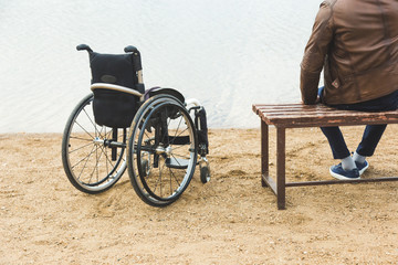 A young man sits on a bench by the lake, next to his wheelchair.