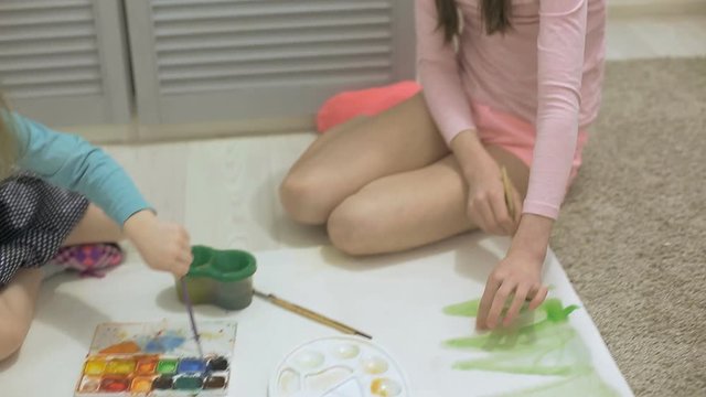 two girls paint with watercolor paints and brushes on a large drawing paper on the floor , children's joint creativity