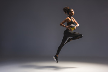 side view of athletic african american sportswoman holding ball and jumping on grey