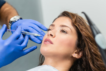 Obraz na płótnie Canvas Attractive young woman is getting a rejuvenating facial injections. She is sitting calmly at clinic. The expert beautician is filling female wrinkles by hyaluronic acid.