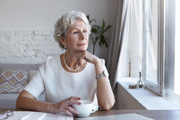 Attractive classy sixty year old mature woman wearing stylish wrist watch and pearl necklace spending nice time at home alone, sitting at her workplace with cup of coffee, looking through window - Powered by Adobe