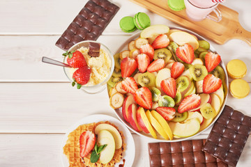 Chocolate bars and fruit sliced ​​from strawberries, apples, bananas and kiwi on a wooden white background.