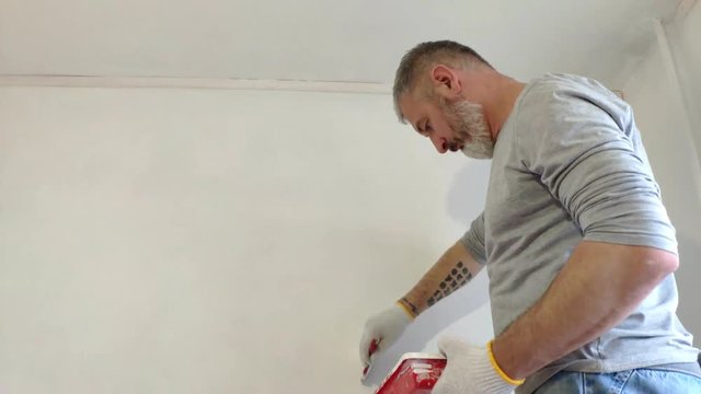 4K Bearded Man Painting interior Walls at Flat using Paint brush. Handsome young man doing Repairs - actively paints wall with white paint. Home renovation or Redecoration concept.