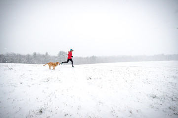 Athletic woman running in field in early season snowstorm with yellow lab shot in profile