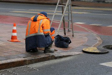 Worker in uniform bent over the open sewer hatch on the sidewalk. Repair of sewage or underground...