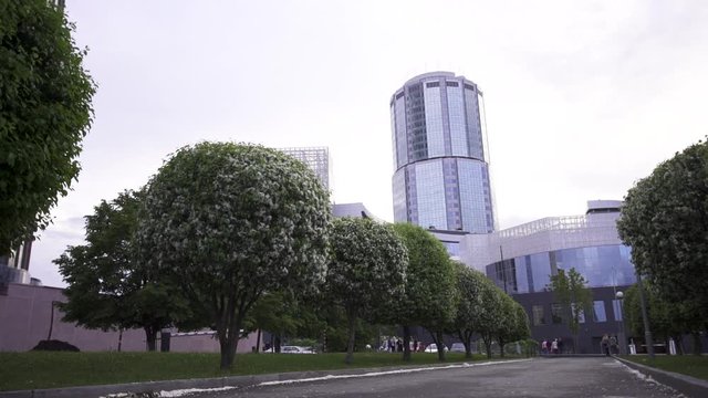 Beautiful spring cityscape with modern buildings standing behing apple bushes. Stock footage. Blossoming apple trees against the background of glass facade buildings.