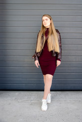 Portrait of a beautiful girl in a dark red dress knee-deep and in a dark leather jacket in the street in the afternoon