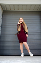 Portrait of a beautiful girl in a dark red dress knee-deep and in a dark leather jacket in the street in the afternoon