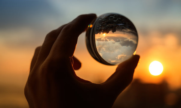 Hand holds glass ball which reflects sunset sky over city.