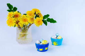 Festive Easter arrangement: dandelion bouquet and decorative painted eggs in ceramic dishes, white background, copy space.