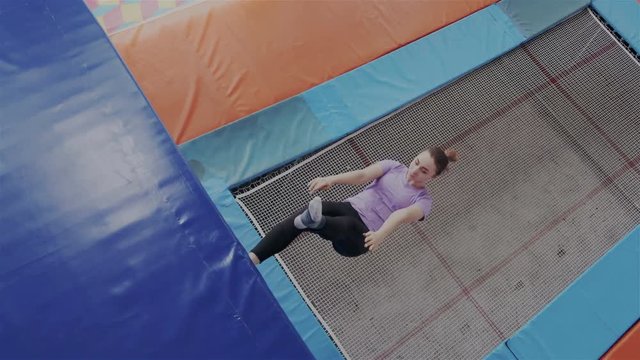 The girl performs sport exercises on a trampoline. She lands on her back, pushes off from the trampoline and takes a few steps along a vertical wall. The girl is happy and joyful