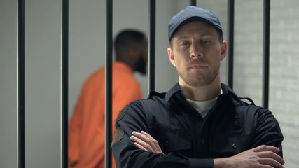 Prison warden looking to camera standing near cell with imprisoned afro-american