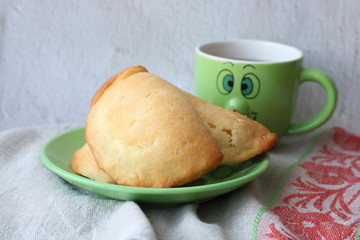 homemade baked cake with cottage cheese lying on a green plate with cup of tea behind it on linen. Russian dish sochni