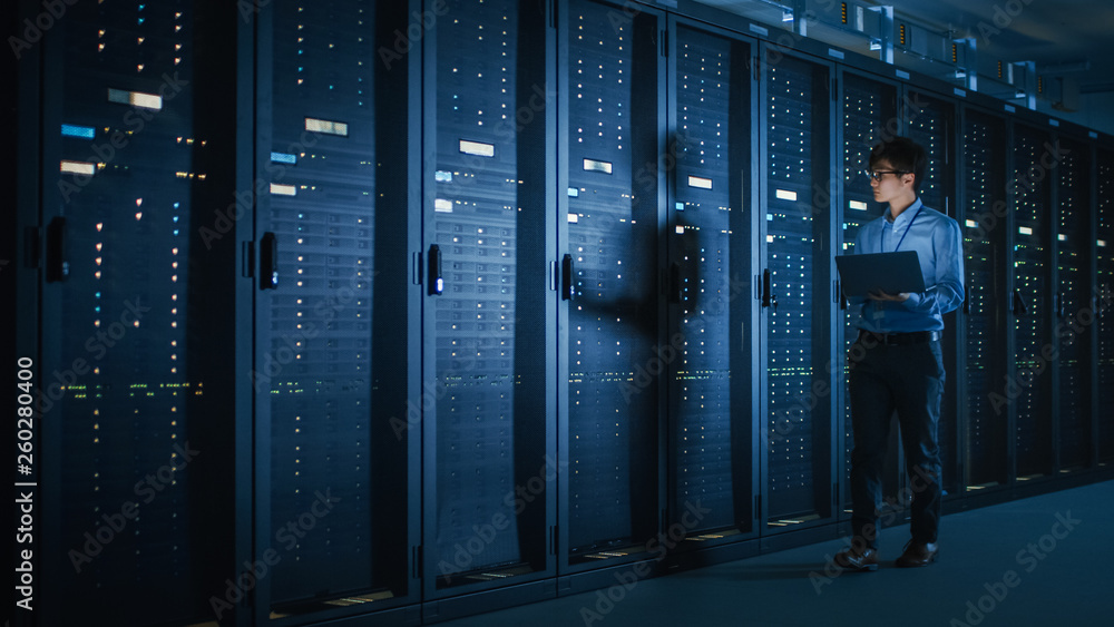 Leinwandbilder in dark data center: male it specialist walks along the row of operational server racks, uses laptop