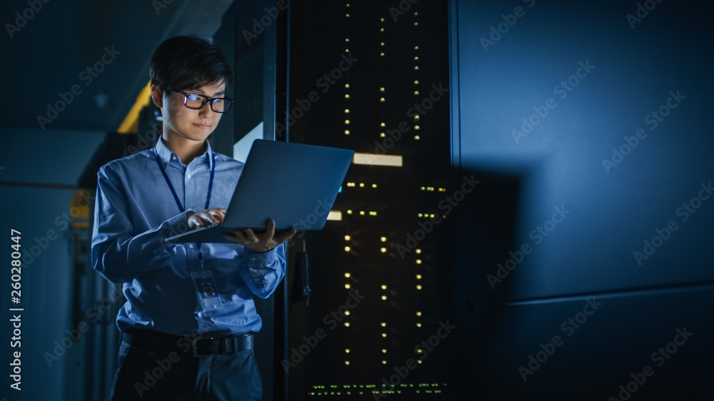 Poster in dark data center: male it specialist stands beside the row of operational server racks, uses lapt