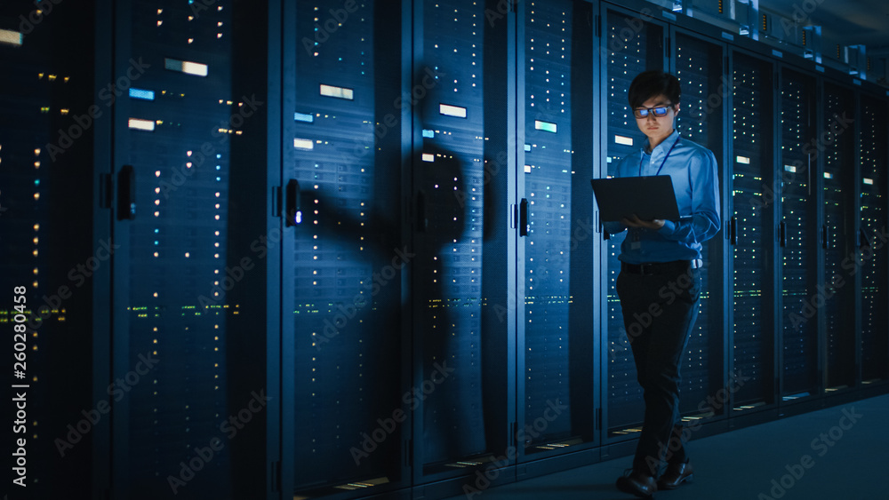 Poster in dark data center: male it specialist walks along the row of operational server racks, uses laptop