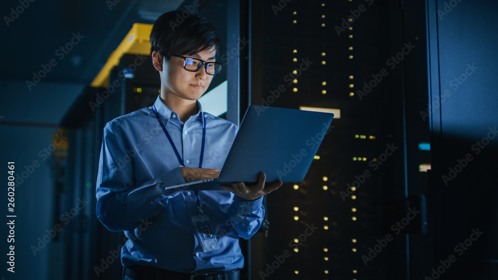 Leinwandbilder in dark data center: male it specialist stands beside the row of operational server racks, uses lapt