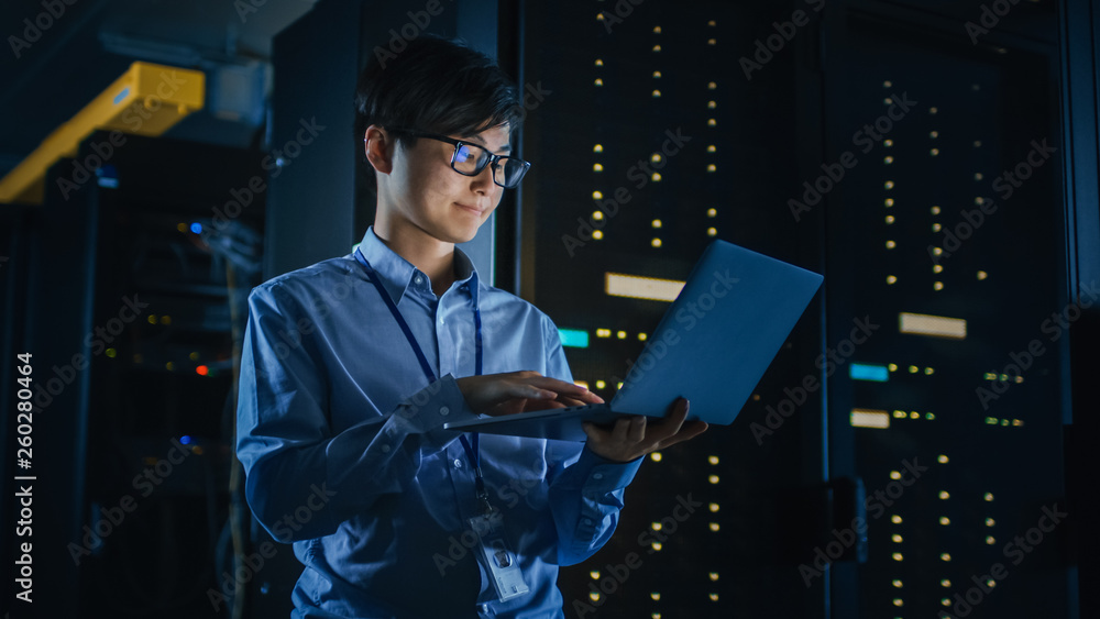 Leinwandbilder in dark data center: male it specialist stands beside the row of operational server racks, uses lapt
