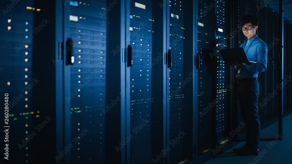 Poster in dark data center: male it specialist stands beside the row of operational server racks, uses lapt