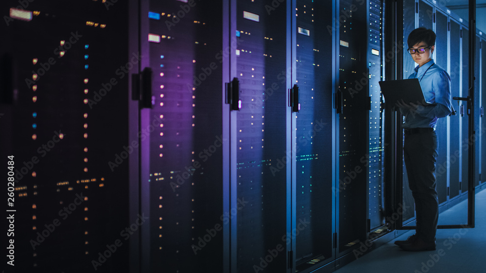 Poster in dark data center: male it specialist stands beside the row of operational server racks, uses lapt