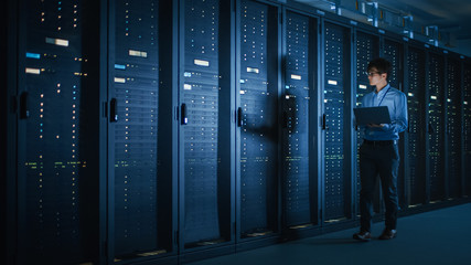 In Dark Data Center: Male IT Specialist Walks along the Row of Operational Server Racks, Uses...