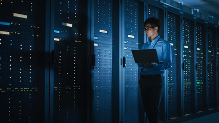 In Dark Data Center: Male IT Specialist Walks along the Row of Operational Server Racks, Uses...