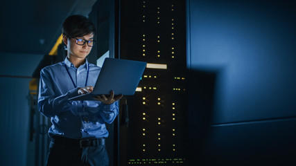 In Dark Data Center: Male IT Specialist Stands Beside the Row of Operational Server Racks, Uses...