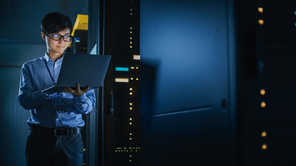 In Dark Data Center: Male IT Specialist Stands Beside the Row of Operational Server Racks, Uses Laptop for Maintenance. Concept for Cloud Computing, Artificial Intelligence, Cybersecurity. Neon Lights