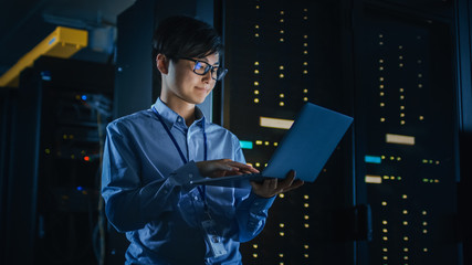 In Dark Data Center: Male IT Specialist Stands Beside the Row of Operational Server Racks, Uses...