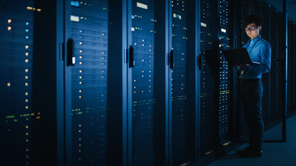 In Dark Data Center: Male IT Specialist Stands Beside the Row of Operational Server Racks, Uses...