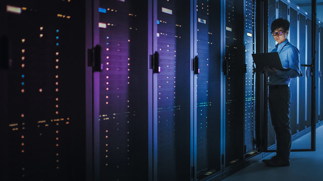 In Dark Data Center: Male IT Specialist Stands Beside the Row of Operational Server Racks, Uses Laptop for Maintenance. Concept for  Artificial Intelligence, Supercomputer, Cybersecurity. Neon Lights