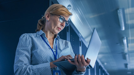 In Data Center: Low-Angle Portrait Shot of a Female IT Technician Running Maintenance Programme on Laptop, Controls Operational Server Rack. Modern High-Tech Telecommunications Operational Data Center