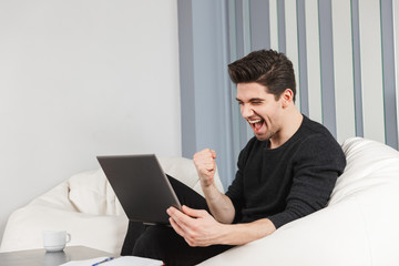Happy handsome young man at home indoors using laptop computer make winner gesture.