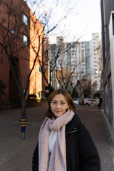 Fototapeta premium Chinese girl standing in a korean street in Jeju island wearing a black jacket and pink scarf ,looking happy