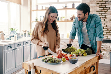 Real love. Beautiful young couple cooking dinner and drinking wine while standing in the kitchen at home