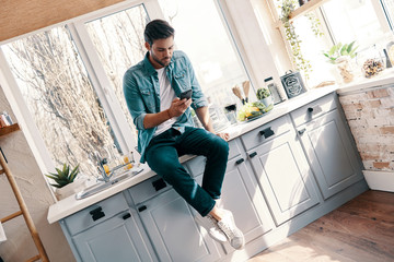 Texting to friends. Handsome young man in casual wear using his smart phone while sitting in the kitchen at home