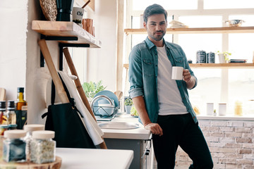 Morning coffee. Handsome young man in casual wear looking at camera and having a hot drink while...