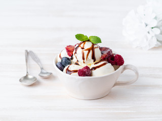 Balls of vanilla ice cream in mug with raspberry and blueberries, chocolate syrup  topping. Side view, white bright background