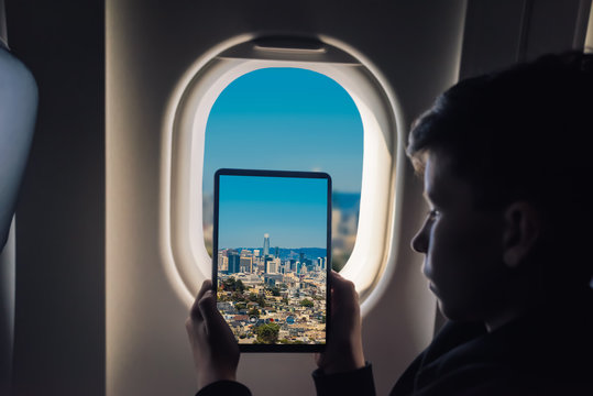 Caucasian boy using tablet pc taking picture through airplane window San Francisco cityscape with skyscrapers. California. USA