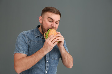 Young man eating tasty burger on grey background. Space for text