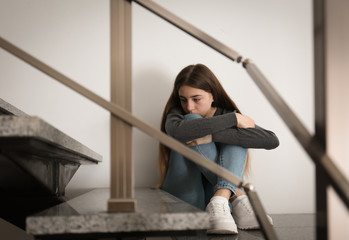 Upset teenage girl sitting on stairs indoors. Space for text