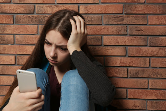Upset teenage girl with smartphone sitting at wall indoors. Space for text