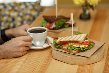man takes a cup of coffee from a wooden table, on which lies a sandwich