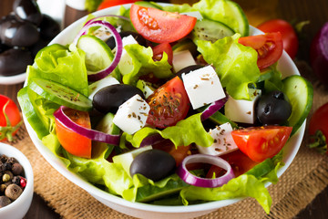 Fresh Greek salad made of cherry tomato, ruccola, arugula, feta, olives, cucumbers, onion and spices. Caesar salad in a white bowl on wooden background. Healthy organic diet food concept.