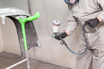 A male worker paints with a spray gun a part of the car body in silver after being damaged at an accident. Bumper from the vehicle during the repair in the workshop.